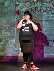 Amy Lamé is standing infront of a brick wall which has a cropped part of the Open Barbers logo projected on it. Amy is wearing red converse trainers, black socks, black leggings, a black skirt and a black tshirt which says #stand by your trans in white. Amy is speaking into a microphone. Amy has black hair and glasses.