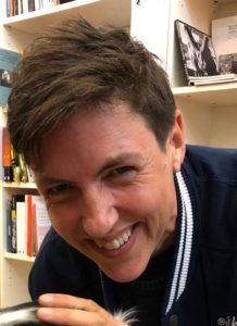 Meg-John Barkers is leaning forward infront of a bookshelf. Meg-John has short brown hair and is smiling. Meg-John is wearing a navy blue top with white stripes at the collar.
