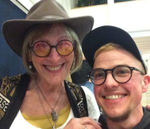 Kate Bornstein is standing next to Greygory Vass with a hand on Greygory's shoulder. They are in a room that has blue and white interior. Kate is wearing a brown hat and glasses with a tint that fades from pink to yellow. Katie is wearing two necklaces and a white top with a yellow top underneath, and a brown, black and white patterned waistcoat. Kate has blonde bobbed hair with a fringe and is smiling. Greygory is wearing a blue baseball cap and has short blonde hair and dark rimmed glasses. Greygory is smiling and has a bearded chin and a dark top.