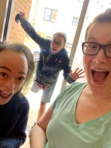 Liz Carr (left) and Jo Church (right) are in the foreground and Greygory Vass is falling through the open door in the background. Liz has dark hair and is wearing a navy blue top. Jo has short, brown hair, is wearing a grean v neck t-shirt and dark rimmed glasses. Both Liz and Jo have open mouths and excited expression. Greygory is wearing a blue denim jacket and short and a grey beany. Greygory has dark rimmed glasses and an open mouth with shocked expression. Greygory is standing on one leg and has arms outretched as if falling or flying. There is a white building in the background with windows.