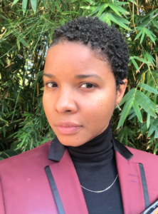Madin Lopez is in front of a green plant. Madin is wearing a purple jacket, black polo neck jumper and silver chain. Madin has short, black curly hair. Madin is looking at the camera with a sincere expression.