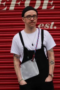 Felix from Open Barbers is standing in front of a red shop front shutter which has some white lettering which is partially cropped from view. Felix is modelling a bag with a red and blue shoulder strap made by Marlo Mortimer as a crowdfunder reward. Felix is wearing a white t-shirt, black dungarees and a black beany hat. He has a moustache and round dark glasses. He has a brown wrist watch and tattoos on his forearms.