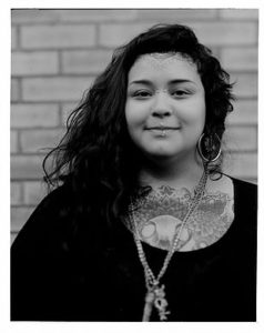 The image is monochrome. Mercedez is standing in front of a brick wall. Mercedez is wearing a black top and a long necklace. Mercedez hs long dark hair and is smiling at the camera. Mercedez has one visible earring, a lip piercing, and a piercing at the bridge of the nose. Mercedez has a tattoos on the chest and forehead.