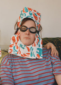 The artists Amy Pennington is sitting wearing swimming goggles which hold and orange and blue print around their Amy's face. A hole is cut out of the print which Amy's face fits through. Amy has a sincere expression, and is wearing an orange and blue striped t-shirt. There is a hand on Amy's shoulder to the right of the photo.